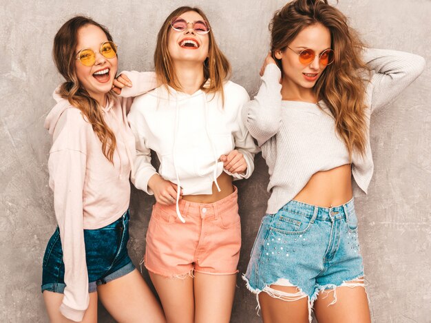 Tre giovani belle ragazze sorridenti in abiti sportivi alla moda estate. Posa sexy spensierata delle donne. Modelli positivi in occhiali da sole rotondi che si divertono