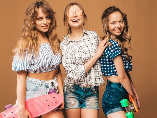 Tre giovani belle ragazze sorridenti sexy alla moda con i pattini variopinti del penny. Le donne in estate camicia a scacchi vestiti in posa in occhiali da sole. Modelli positivi che si divertono