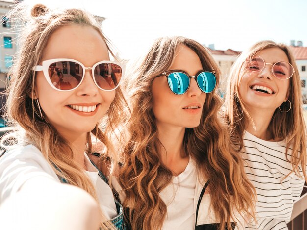 Tre giovani donne sorridenti dei pantaloni a vita bassa in vestiti di estate Ragazze che prendono le foto dell'autoritratto del selfie sullo smartphone Modelli che posano nella via Femmina che mostra le emozioni positive del fronte
