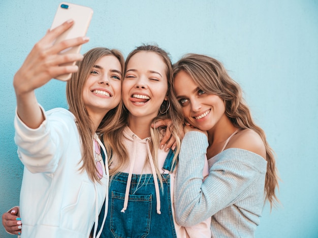 Tre giovani donne sorridenti dei pantaloni a vita bassa in vestiti di estate Ragazze che prendono le foto dell'autoritratto del selfie sullo smartphone Modelli che posano nella via vicino alla parete Femmina che mostra le emozioni positive del fronte Lingua di mostra