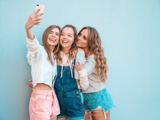 Tre giovani donne sorridenti dei pantaloni a vita bassa in vestiti di estate Ragazze che prendono le foto dell'autoritratto del selfie sullo smartphone Modelli che posano nella via vicino alla parete Femmina che mostra le emozioni positive del fronte Lingua di spettacoli