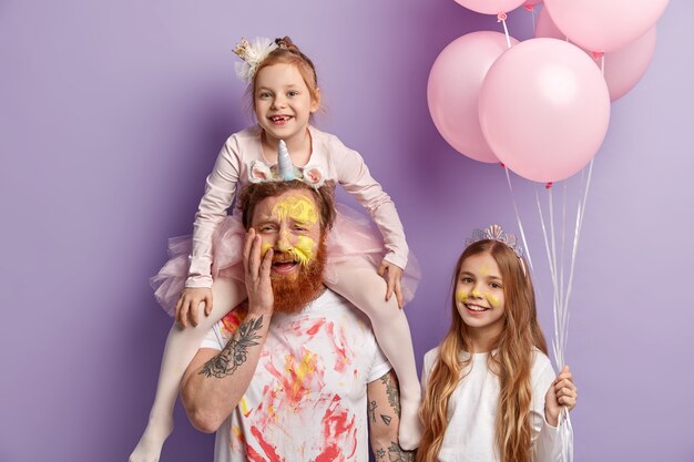 Tre membri della famiglia pongono al coperto sopra la parete viola. Divertenti due figlie e papà si divertono, colorano i volti con acquerelli colorati, festeggiano insieme la festa dei bambini. Concetto di intrattenimento.