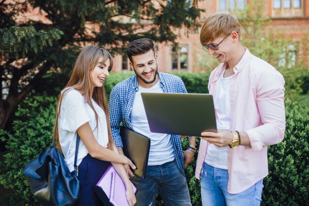 Tre studenti in abiti eleganti guardano un laptop e ridono del campus universitario.