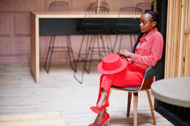 Trecce graziose affari afroamericani signora brillante persona prepotente amichevole indossare ufficio camicia rossa cappello e pantaloni sedersi sulla sedia
