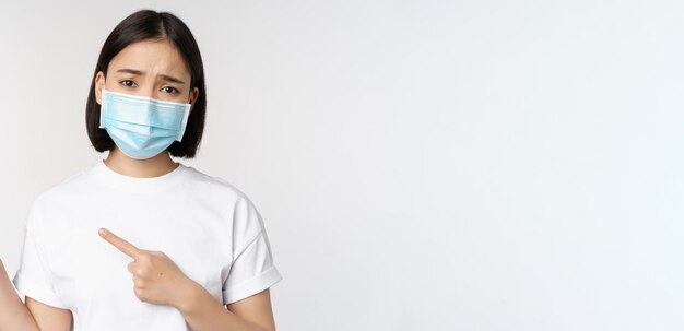 Triste donna asiatica in maschera medica che punta le dita a sinistra accigliata e sembra sconvolta lamentandosi dimostrando banner in piedi su sfondo bianco