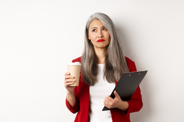 Triste donna d'affari asiatica che beve caffè al lavoro e guarda l'angolo in alto a sinistra, in piedi su sfondo bianco