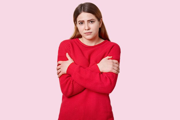 Triste donna dai capelli scuri incrocia le mani sul petto, ha il trucco, vestita in maglione rosso sciolto, modelle sul rosa