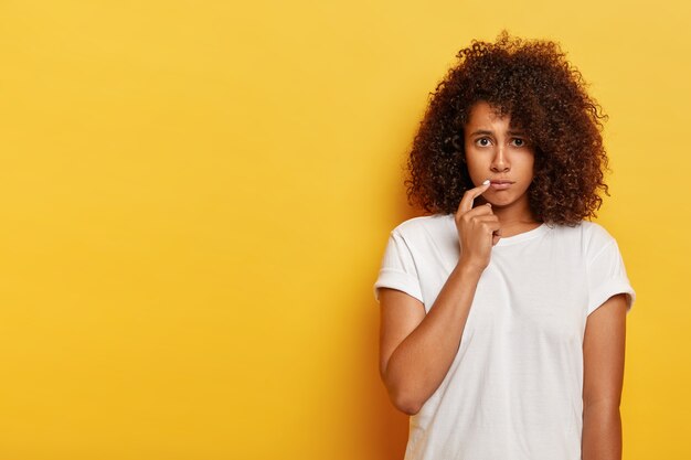 Triste donna millenaria afroamericana imbronciata, tiene il dito indice vicino al cereale delle labbra, insultata da qualcuno, si sente delusa, vestita in abbigliamento casual, posa contro il muro giallo, ha problemi