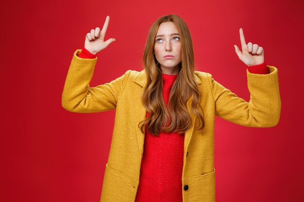 Triste e triste donna rossa carina in cappotto giallo alzando le mani scontento di rimpianto guardando l'angolo in alto a sinistra apatico come deluso dalla pioggia o dal maltempo sul muro rosso.