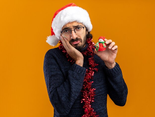 Triste giovane bel ragazzo che indossa il cappello di Natale con la ghirlanda sul collo tenendo il giocattolo mettendo la mano sulla guancia isolato su sfondo arancione