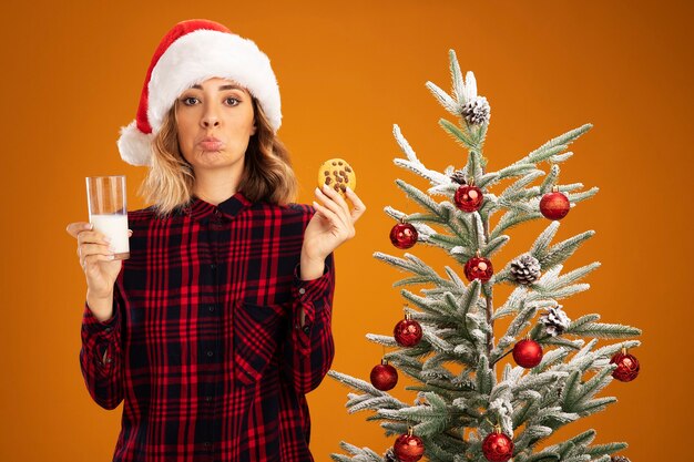 Triste giovane bella ragazza in piedi vicino all'albero di Natale con cappello di Natale che tiene in mano un bicchiere di latte con biscotti isolati su sfondo arancione
