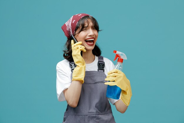 Triste giovane donna addetta alle pulizie che indossa bandana uniforme e guanti di gomma che tengono il detergente parlando al telefono guardando il lato isolato su sfondo blu