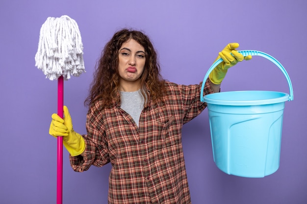 Triste giovane donna delle pulizie che indossa guanti che tengono mop con secchio isolato su parete viola