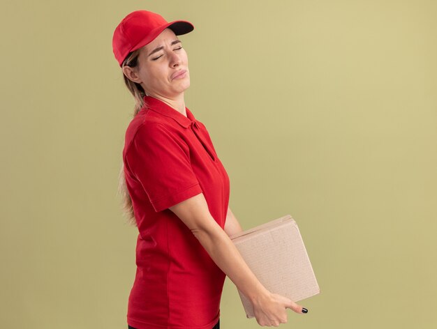 Triste giovane donna graziosa di consegna in uniforme si leva in piedi lateralmente tenendo cardbox isolato sulla parete verde oliva