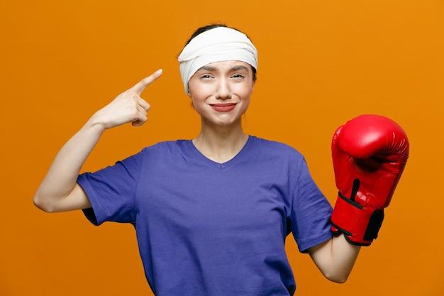 Triste giovane donna sportiva che indossa maglietta e guantoni da boxe guardando la fotocamera tenendo la mano in aria puntando verso la testa con la testa avvolta con benda isolata su sfondo arancione