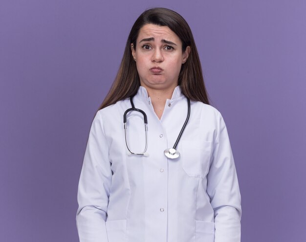 Triste giovane dottoressa che indossa un abito medico con lo stetoscopio che soffia sulle guance isolate sulla parete viola con spazio di copia