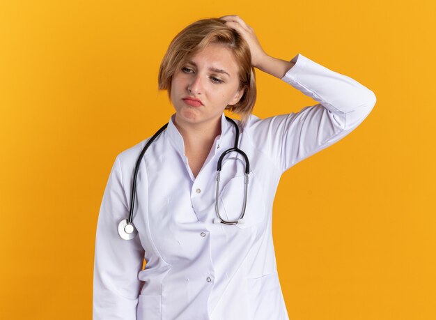 Triste giovane dottoressa che indossa una tunica medica con uno stetoscopio che mette la mano sulla testa isolata su sfondo arancione