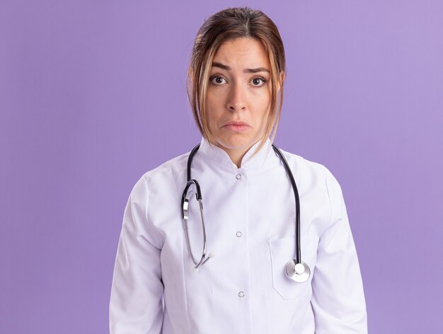 Triste giovane medico femminile che indossa abito medico con lo stetoscopio isolato sulla parete viola