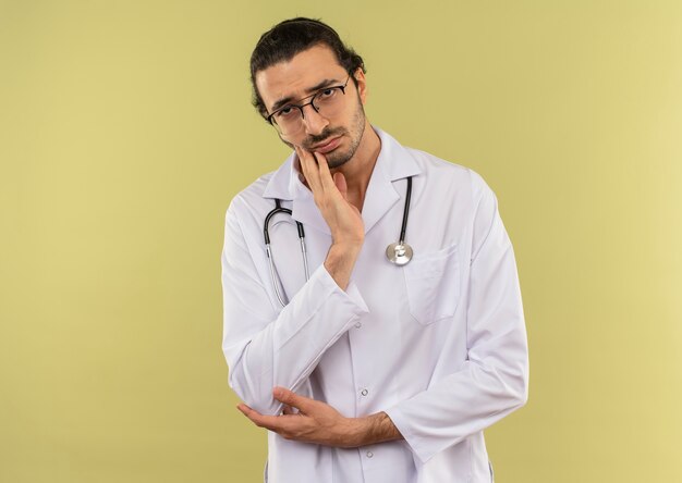 Triste giovane medico maschio con occhiali ottici che indossa una veste bianca con uno stetoscopio che mette la mano sul mento su sfondo verde green