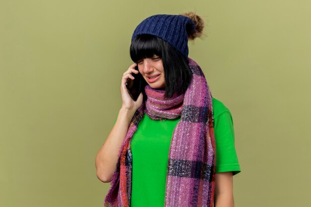 Triste giovane ragazza caucasica malata indossando cappello invernale e sciarpa parlando al telefono guardando verso il basso isolato su sfondo verde oliva con spazio di copia
