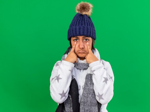 Triste giovane ragazza malata che indossa il cappello invernale con sciarpa mettendo le dita sugli occhi isolati su sfondo verde