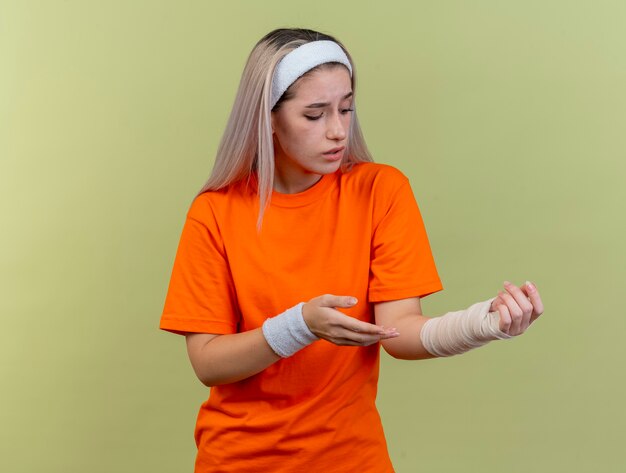 Triste giovane ragazza sportiva caucasica con bretelle che indossa fascia e braccialetti punta e guarda a portata di mano