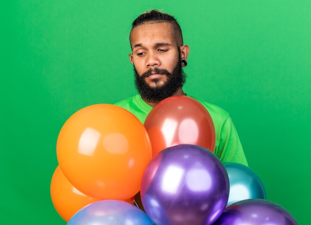 Triste giovane ragazzo afroamericano che indossa una maglietta verde in piedi dietro palloncini isolati sul muro verde