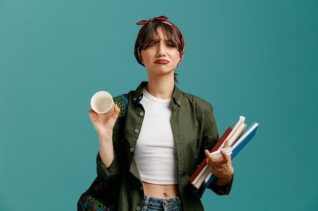 Triste giovane studentessa che indossa bandana e zaino con in mano blocchi di appunti tazza di caffè di carta e il suo cappuccio guardando la fotocamera che mostra una tazza di caffè vuota isolata su sfondo blu