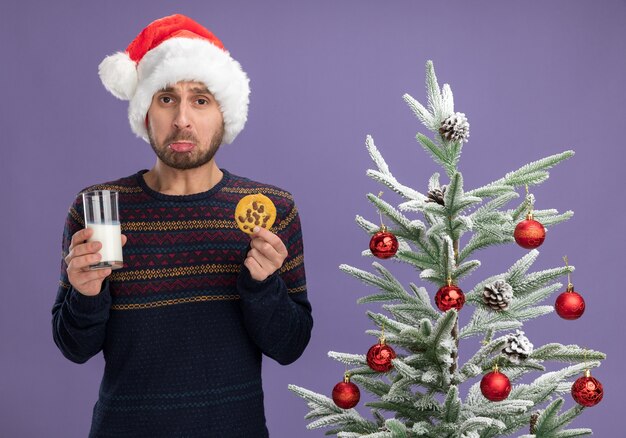 Triste giovane uomo caucasico che indossa il cappello di Natale in piedi vicino all'albero di Natale decorato tenendo un bicchiere di latte e biscotto guardando la telecamera isolata su sfondo viola