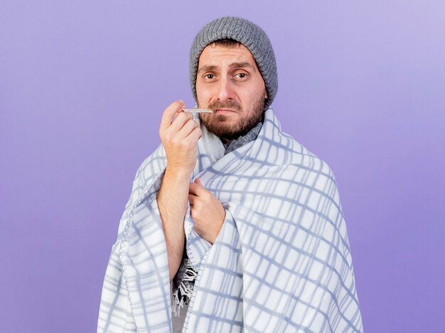 Triste giovane uomo malato che indossa un cappello invernale con sciarpa tenendo il termometro in bocca isolato su sfondo viola