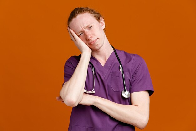 Triste mettendo la mano sulla guancia giovane medico maschio che indossa l'uniforme con lo stetoscopio isolato su sfondo arancione