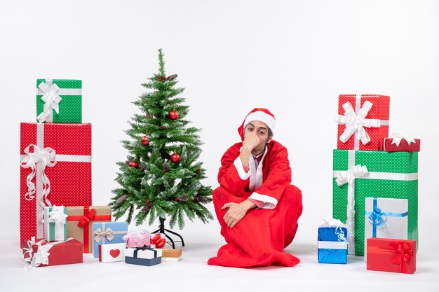 Triste pensieroso giovane uomo vestito da Babbo Natale con doni e albero di Natale decorato seduto per terra su sfondo bianco
