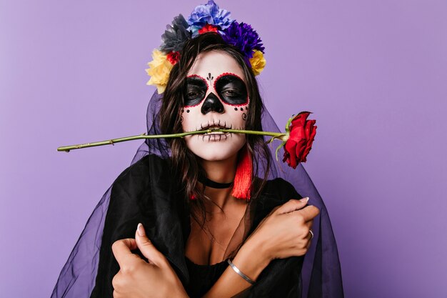 Triste sposa morta in velo nero in posa di halloween. Donna arrabbiata con la pittura messicana del viso in piedi sul muro viola.