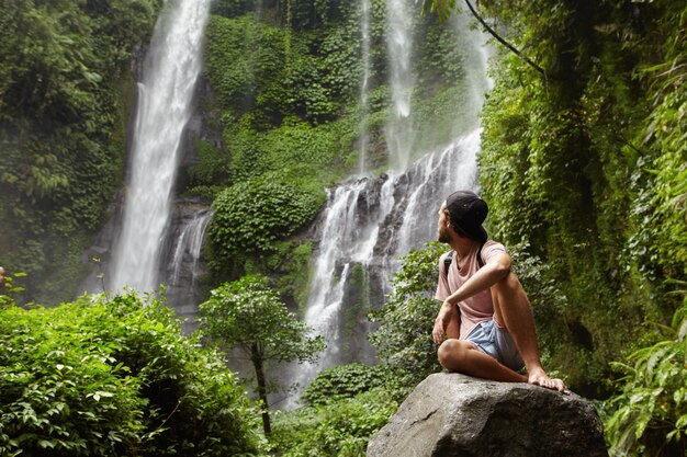 Turismo, viaggi e avventura. Elegante giovane hipster seduto sulla pietra con i piedi nudi e voltando la testa indietro per vedere una cascata incredibile