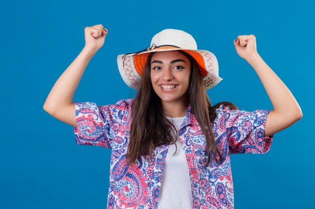 turista donna con cappello che sembra uscito rallegrandosi del suo successo e della vittoria stringendo i pugni con gioia felice di raggiungere il suo scopo e gli obiettivi in piedi sul blu isolato