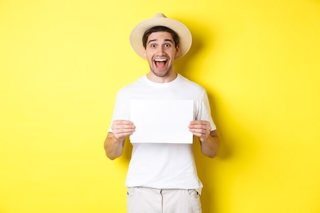 Turista eccitato che mostra il tuo logo o segno su un pezzo di carta bianco, sorridendo stupito, in piedi su sfondo giallo.