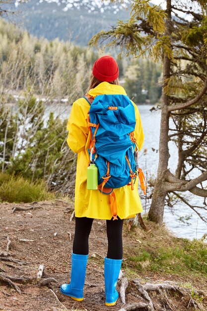Turista femminile si alza alla telecamera, vestito con un impermeabile giallo casual, stivali di gomma, respira aria fresca vicino al lago di montagna, conduce uno stile di vita attivo