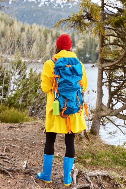 Turista femminile si alza alla telecamera, vestito con un impermeabile giallo casual, stivali di gomma, respira aria fresca vicino al lago di montagna, conduce uno stile di vita attivo