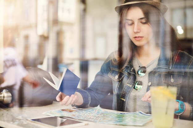 Turista seduto in un caffè a leggere una mappa in possesso di passaporto bevendo limonata in una città frenetica Girato attraverso il vetro