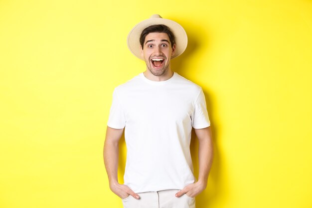 Turista sorpreso in cappello di paglia che sembra felice, reagisce stupito alla pubblicità dell'agenzia di viaggi, in piedi su sfondo giallo.