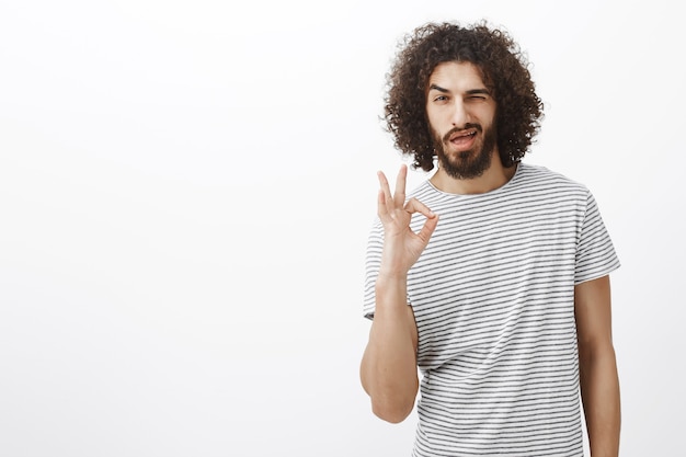 Tutto sotto controllo piccola. Bell'uomo ispanico fiducioso con barba e acconciatura riccia, mostrando segno ok o ok, ammiccante