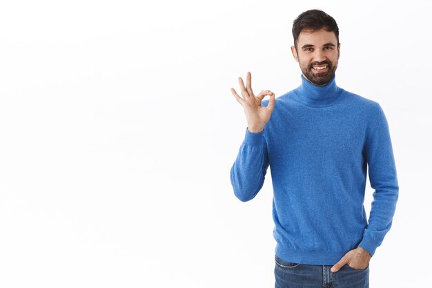 Tutto sotto controllo. Ritratto di successo felice caucasico, imprenditore maschio con la barba, mostra bene, gesto ok e sorridente soddisfatto, consiglia un prodotto di buona qualità, muro bianco