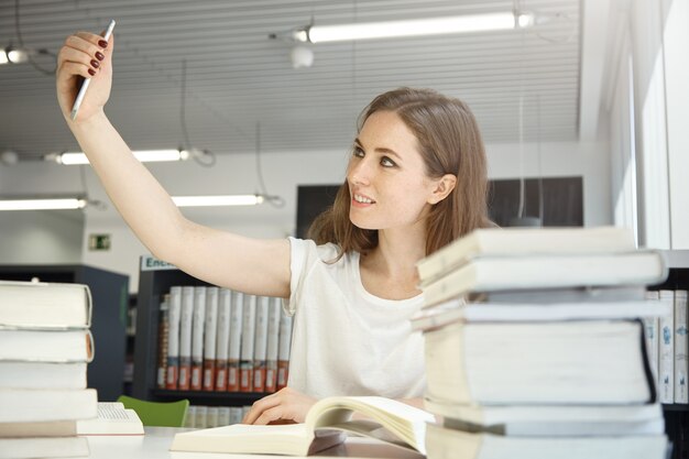 Umano e tecnologia. Persone ed educazione. Ritratto dell'interno della donna adolescente caucasica alla biblioteca che prova a prendere un selfie, circondato da libri e manuali