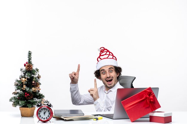 Umore del nuovo anno con il giovane uomo d'affari eccitato sorridente sorpreso che si siede nell'ufficio e che indica sopra su fondo bianco