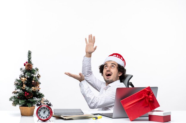 Umore del nuovo anno con il giovane uomo d'affari positivo sorridente con il cappello del Babbo Natale che si siede nell'ufficio e che lavora da solo sul progetto su fondo bianco