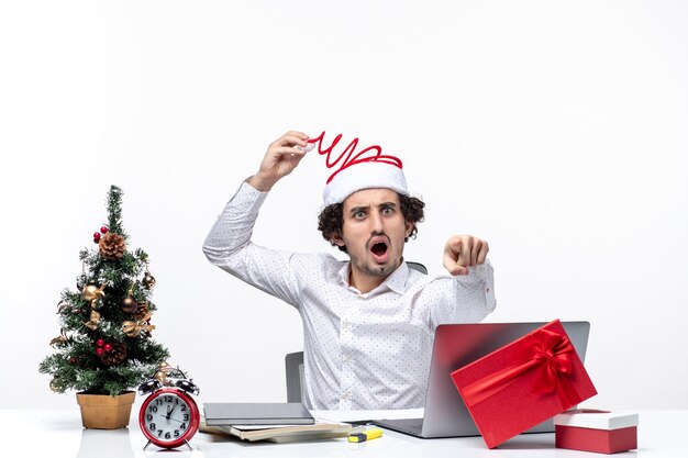 Umore del nuovo anno con il giovane uomo d'affari sorpreso che tocca il suo cappello divertente del Babbo Natale che indica qualcosa in avanti nell'ufficio su fondo bianco