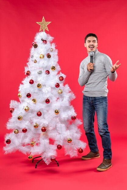 Umore del nuovo anno con il ragazzo positivo che canta la canzone che sta vicino all'albero di Natale decorato sul rosso