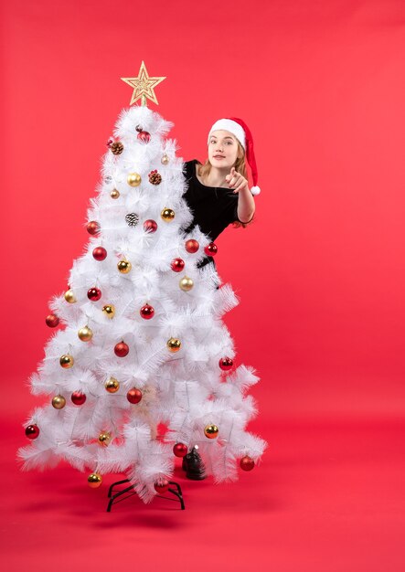 Umore del nuovo anno con la giovane donna in vestito nero e cappello di Babbo Natale in piedi dietro l'albero di Natale bianco