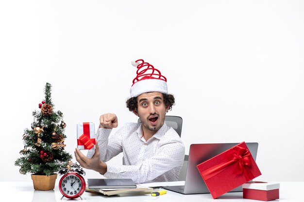 Umore di Natale con il giovane uomo d'affari emotivo divertente con il cappello di Babbo Natale e indicando il suo regalo su priorità bassa bianca