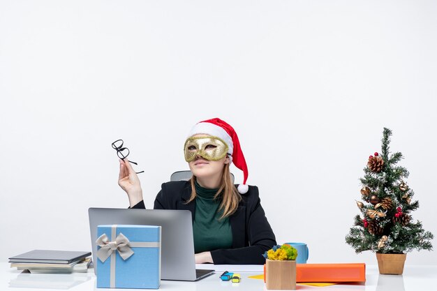 Umore di natale con la giovane donna positiva sognante con il cappello di babbo natale che tiene gli occhiali e la maschera da portare seduto a un tavolo su priorità bassa bianca